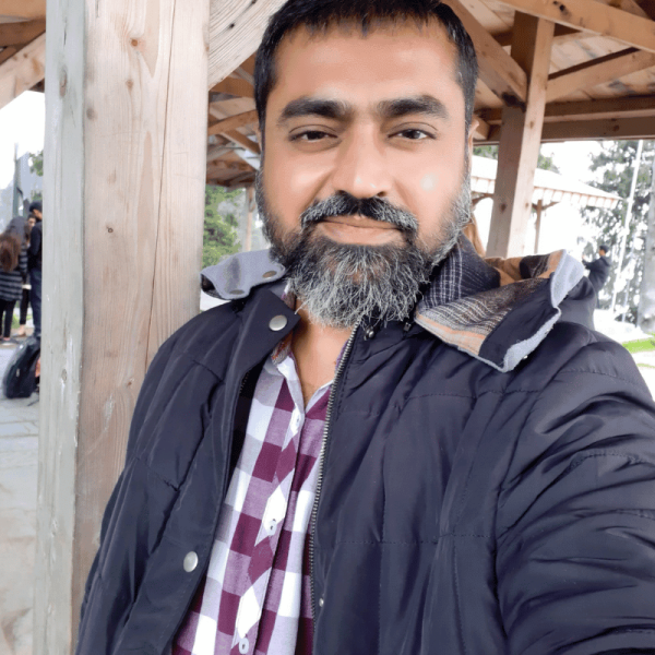 Muhammad Asim outdoors in casual attire, wearing a checked shirt and jacket, smiling at the camera